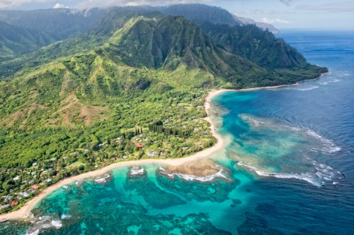 Aerial view of lush green mountains meeting a turquoise ocean, with a sandy beach and coral reefs along the coastline.