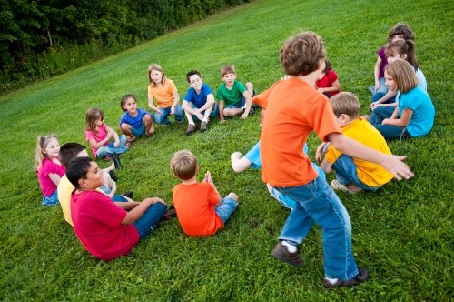 A group of children in colorful shirts sitting in a circle on grass, with one child playfully moving in the center.