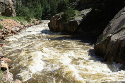 A rushing river flows between rocky banks, surrounded by lush greenery and steep cliffs under a clear blue sky.