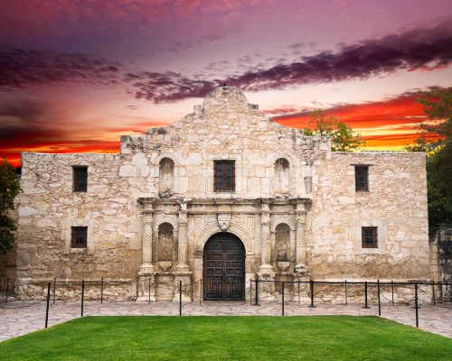 The Alamo, a historic stone mission, set against a vibrant sunset sky. Green grass and a fence in the foreground.