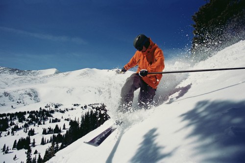 A skier in an orange jacket carves through fresh snow on a mountain slope under a clear blue sky.