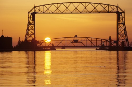 Sunset over a bridge with a silhouette of a lift bridge, reflecting on calm water, with ducks swimming nearby.