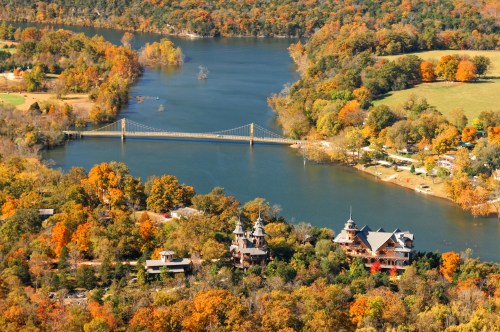 Aerial view of a river surrounded by autumn foliage, featuring a bridge and two distinctive buildings along the shore.
