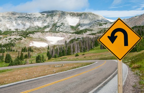 A winding road with a U-turn sign, surrounded by mountains and greenery under a blue sky.