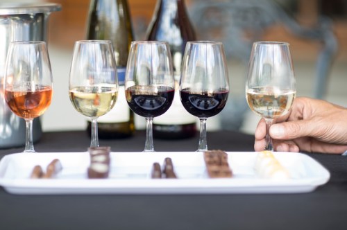 A table with five wine glasses and a platter of assorted chocolates, with a hand holding a glass of white wine.