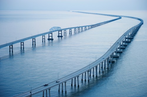 A long, winding bridge stretches over calm waters, connecting distant shores under a soft blue sky.