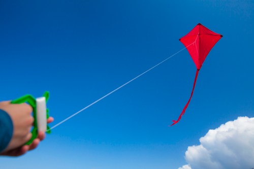 A hand holds a green kite reel, flying a bright red kite against a clear blue sky with fluffy white clouds.