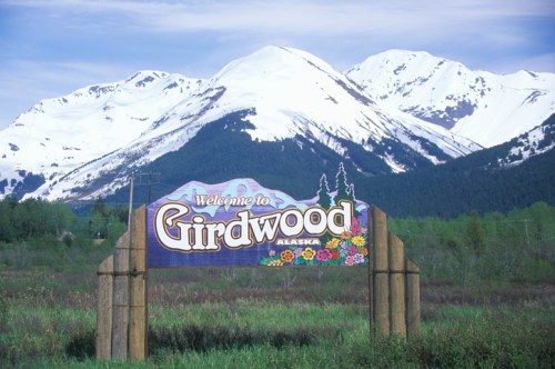 A colorful welcome sign for Girdwood, Alaska, with snow-capped mountains in the background and green fields in the foreground.