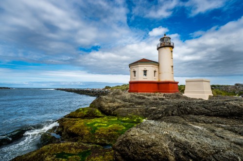 A lighthouse stands on rocky shores, surrounded by water under a cloudy sky. Green moss covers the rocks.