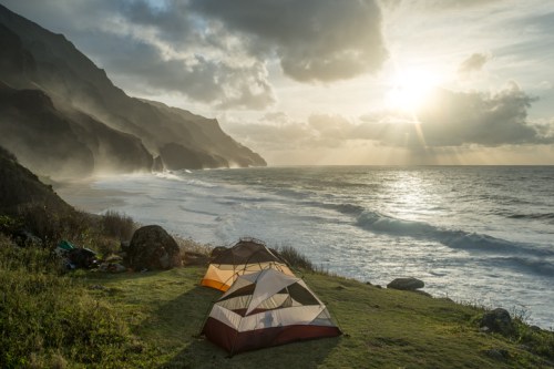 Tents set up on a grassy shore with waves crashing, mountains in the background, and a dramatic sunset sky.