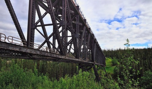 A large metal bridge spans a lush green valley, surrounded by dense trees under a partly cloudy sky.