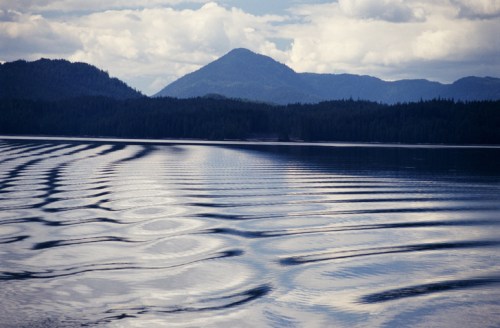 Calm lake with gentle ripples, surrounded by mountains and a cloudy sky.