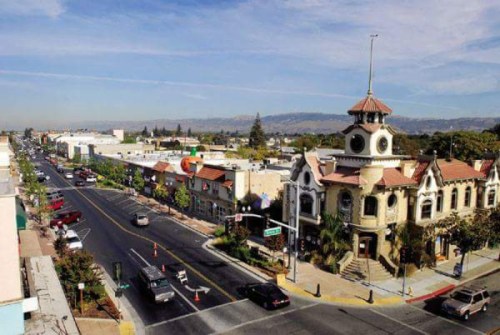 Aerial view of a bustling street with shops and a historic building, surrounded by mountains under a clear blue sky.