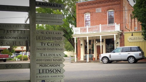 Signpost with winery directions in front of a brick building and parked white SUV on a quiet street.