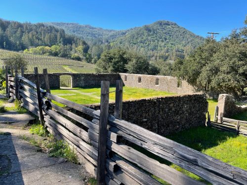 Stone ruins surrounded by a wooden fence, with rolling hills and vineyards in the background under a clear blue sky.