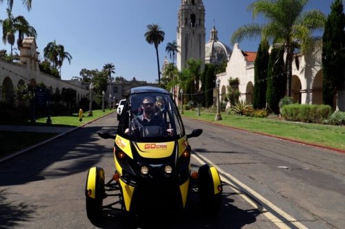 A person drives a yellow and black GoCar on a sunny street lined with palm trees and historic buildings.