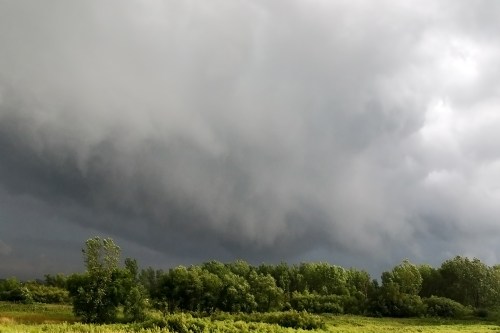 Dark, ominous storm clouds loom over a green landscape with trees, suggesting an approaching storm.
