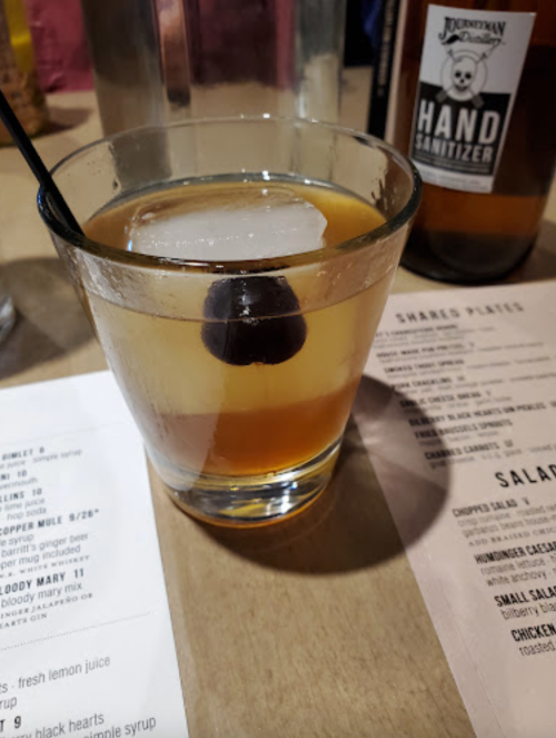 A cocktail with ice and a cherry sits on a table next to a menu and a bottle of hand sanitizer.
