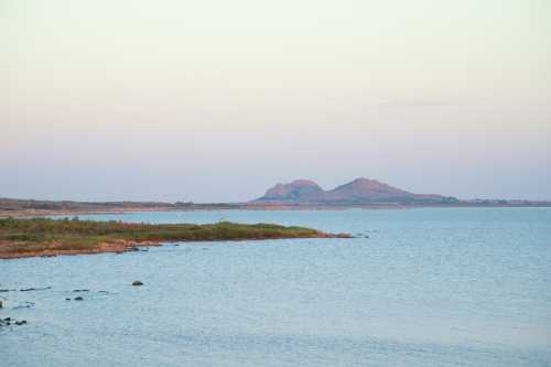 A serene view of a calm sea with distant hills under a pastel sky at dusk.