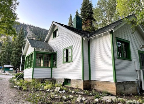 A charming white house with green trim, surrounded by trees and mountains, featuring large windows and a sloped roof.
