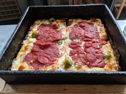 A square pan of cheesy pizza topped with slices of pepperoni and green olives, baked to a golden brown.