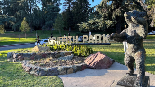 Griffith Park entrance with a bear statue and visitors walking in a green, tree-filled area.