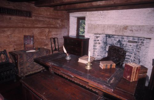 A rustic room with wooden walls, a large table, chairs, a fireplace, and books on a shelf, evoking a historical atmosphere.