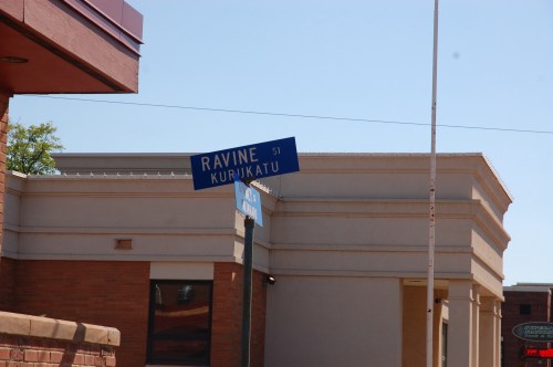 Street sign for Ravine Street and Kurikatu, with a building in the background under a clear blue sky.