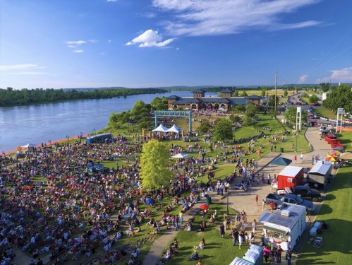 A large crowd gathers at a riverside park for an outdoor event, with food trucks and a stage in the background.