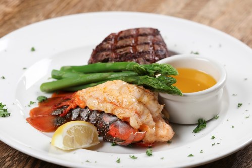 A plate featuring grilled steak, lobster tail, asparagus, and a lemon wedge, served with a small bowl of sauce.