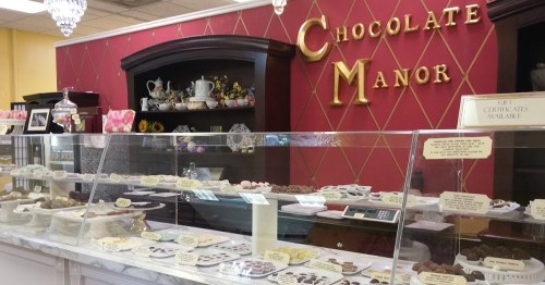 A display case filled with various chocolates in a shop, with a red wall and decorative items in the background.