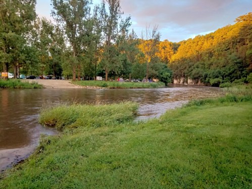 A serene river scene with lush green grass, trees, and a sandy bank under a colorful sunset sky.