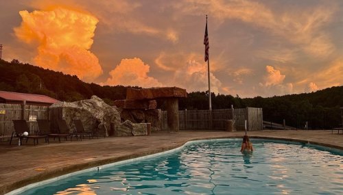 A person wades in a pool at sunset, with vibrant clouds and an American flag in the background.