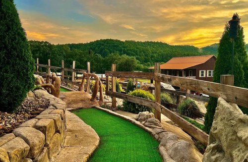 A scenic mini-golf course with a green path, wooden fences, and a sunset over rolling hills in the background.