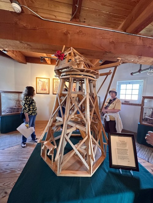 A wooden model structure with intricate designs, displayed in a room with visitors and informational plaques.