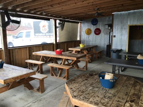 A rustic outdoor dining area with wooden tables, fans, and colorful buckets, featuring a truck visible outside.