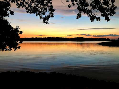 A serene sunset over a calm lake, framed by tree branches, with vibrant colors reflecting on the water's surface.