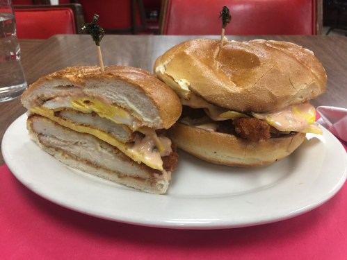 A plate with two sandwiches: one cut in half, featuring chicken, egg, and sauce, and the other whole with similar ingredients.