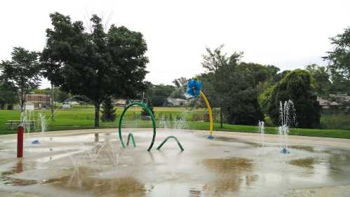 A splash pad with colorful water features, surrounded by green grass and trees under a cloudy sky.