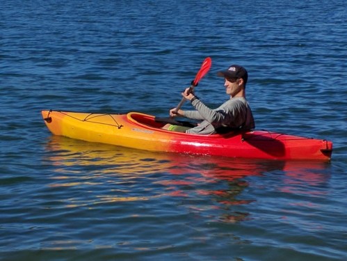 A person paddles a bright orange and red kayak on calm blue water, wearing a cap and a long-sleeve shirt.