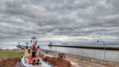 A tugboat in the foreground with a cloudy sky, pier, and people along the waterfront in the background.