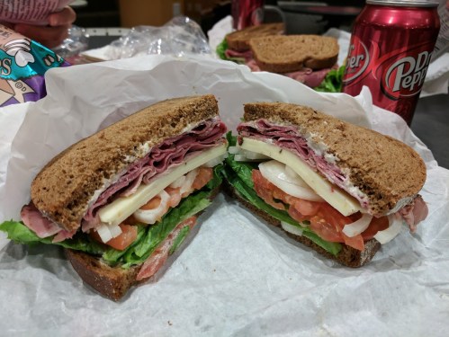 A sandwich with layers of deli meats, cheese, lettuce, and tomato, served on whole grain bread with a can of Dr Pepper.