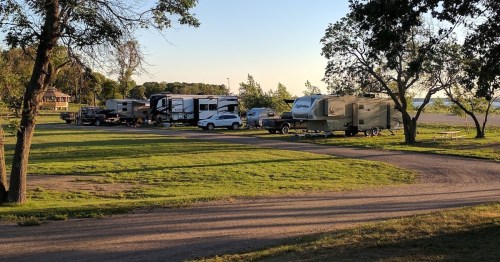 A row of RVs parked in a grassy area near a lake, with trees and a clear sky in the background.
