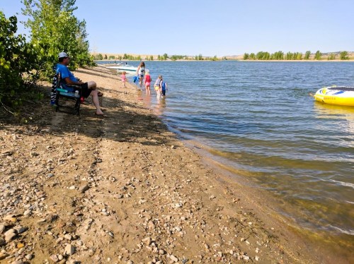 A sandy beach by a river with children playing in the water and an adult sitting on the shore.