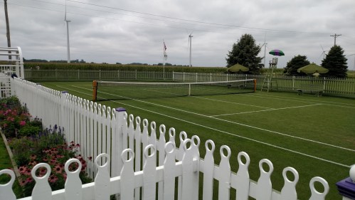A well-maintained tennis court surrounded by a white picket fence, with wind turbines and trees in the background.