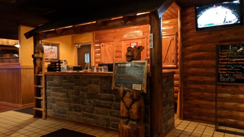 Rustic restaurant entrance with wooden decor, stone accents, and a TV displaying a sports game in the background.