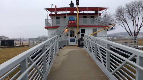 A view of a boat-shaped building with a ramp, featuring a sign that says "Welcome" and "Open." Gray, overcast sky.