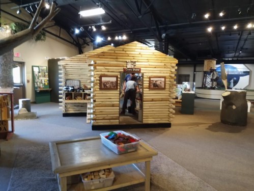 A log cabin exhibit inside a museum, with a person entering and various displays in the background.