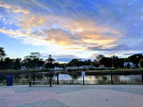 A serene lakeside view at sunset, with colorful clouds reflecting on the water and a paved area in the foreground.