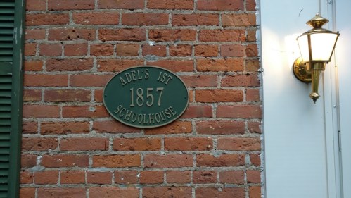 Plaque on a brick wall reading "Adel's 1st Schoolhouse 1857," next to a wall-mounted light fixture.